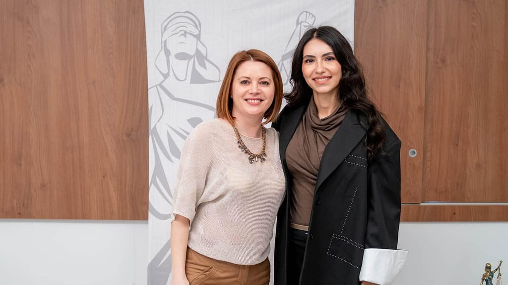 Bianca Opriș stă lângă Ramona Leahu, zâmbind amandoua camerei. Femeia din stânga are părul roșu până la umeri, poartă un top bej cu mâneci scurte și un colier. Femeia din dreapta are părul lung și închis la culoare, poartă o jachetă închisă la culoare peste un top maro. Un banner cu o figură a Lady Justice este pe fundal.