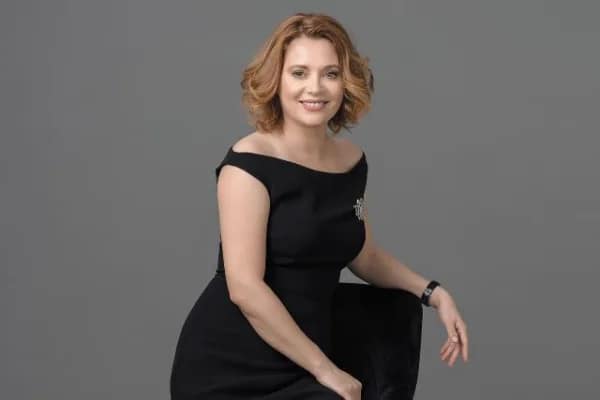 Binaca Opriș cu părul ondulat până la umeri stă pe un scaun negru. Ei poartă o rochie neagră fără umeri și un ceas la încheietura mâinii stângi, zâmbind și pozând pe un fundal gri simplu.